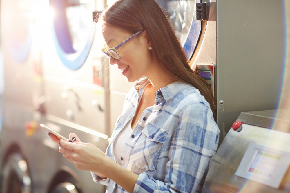 A female managing laundry business using cloud-based software, showing a manager accessing real-time data on a mobile, with laundry machines in the background.
